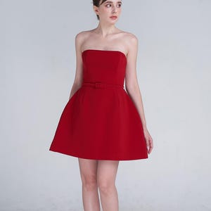 May include: A vibrant red strapless mini dress featuring a fitted bodice and a flared skirt. The dress includes a matching thin belt with a rectangular buckle at the waist, creating a structured silhouette that ends above the knee.