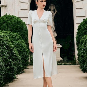 May include: A woman wearing a white midi dress with short sleeves and a button-down front. The dress has a V-neckline and a slit in the front. She is wearing white heels. The background features a stone structure and lush greenery.