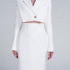 Puede incluir: Un traje blanco de dos piezas con una chaqueta corta con un solo botón dorado y una falda lápiz de cintura alta. La chaqueta tiene mangas largas y solapas con muescas. La falda es entallada y llega a las rodillas. El traje está hecho de una tela lisa y opaca.