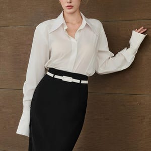May include: A woman's outfit featuring a white button-down shirt with long sleeves and a black pencil skirt. A white belt cinches the waist. The outfit is completed with sunglasses.