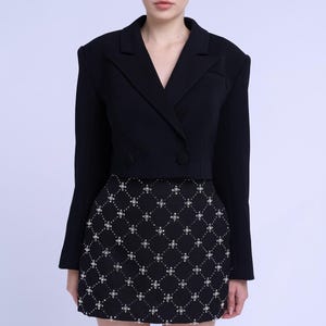 Puede incluir: Una blazer negra con cierre de doble botonadura y cuello en V. La modelo lleva una minifalda negra con un estampado geométrico blanco. El conjunto es elegante y adecuado para diversas ocasiones.