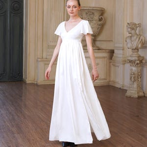 Peut inclure: Une robe blanche longue jusqu'au sol avec un décolleté en V, des manches courtes évasées et un corsage taille haute avec une jupe fluide. Le vêtement est accessoirisé avec un collier de perles simple et un bracelet assorti, offrant un style épuré.