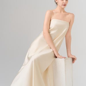 May include: A floor-length, cream-colored satin dress featuring a straight-across neckline and thin spaghetti straps. The garment has a smooth, lustrous finish and a fluid, A-line silhouette that drapes down to the floor in a simple, clean design.