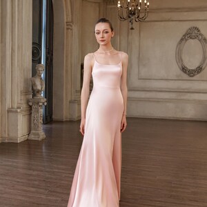 May include: A floor-length, pale pink satin dress featuring a scoop neckline, thin spaghetti straps, and a fitted bodice that transitions into a flowing, A-line skirt. The fabric has a smooth, lustrous finish that reflects light across the surface.
