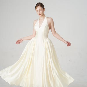 May include: A cream-colored halter-neck gown with a full skirt. The dress features a V-neckline and a flowing, floor-length design. The model is wearing black heels. The dress is suitable for formal events.