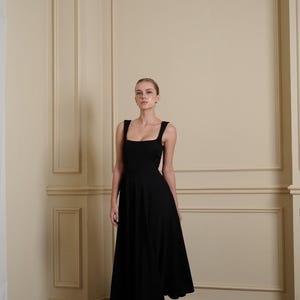 May include: A solid black midi-length dress featuring a square neckline, wide shoulder straps, and a fitted bodice that transitions into a flared, A-line skirt. The garment is constructed from a smooth, opaque fabric and falls below the knees.
