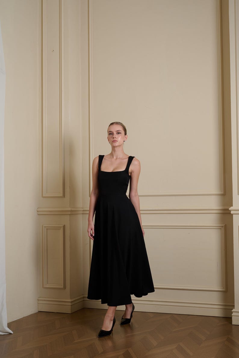 May include: A solid black midi-length dress featuring a square neckline, wide shoulder straps, and a fitted bodice that transitions into a flared, A-line skirt. The garment is constructed from a smooth, opaque fabric and falls below the knees.
