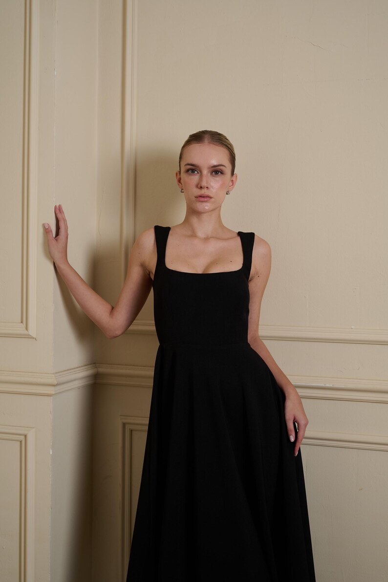 May include: A solid black sleeveless dress featuring a square neckline and wide shoulder straps. The bodice is fitted, transitioning into a full, flowing skirt that extends downward. The garment is constructed from a smooth, opaque fabric.