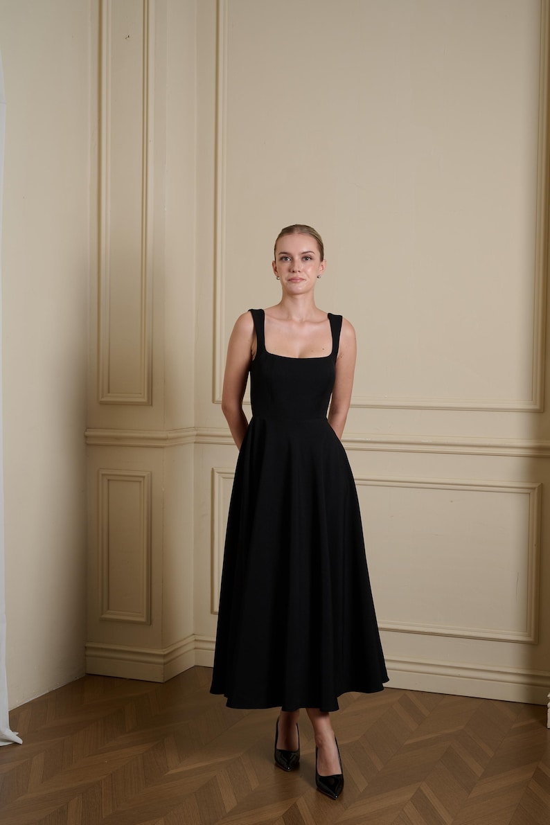 May include: A solid black midi-length dress featuring a square neckline, wide shoulder straps, and a fitted bodice that transitions into a full, flared skirt. The garment is constructed with a clean, structured silhouette and a smooth, matte finish.