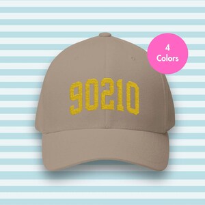 Può includere: Un cappellino da baseball beige con il numero "90210" ricamato in giallo. Un cerchio rosa in alto a destra indica "4 colori" disponibili. Il cappellino è posto su uno sfondo a righe bianche e azzurre.