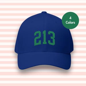 May include: A blue baseball cap with the number "213" embroidered in green on the front. A green circle with the text "4 Colors" is in the upper right corner. The hat is set against a pink and white striped background.