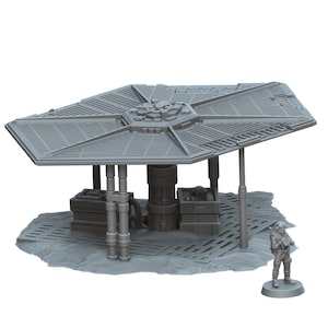 May include: A gray, miniature sci-fi structure with a large, geometric canopy supported by cylindrical pillars. The model includes crates, a central tower, and a small figure of a person in a spacesuit. The base is textured to resemble a rocky terrain.