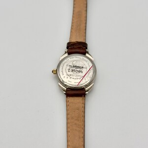 May include: A silver-toned wristwatch with a brown leather band. The watch face is white with black text and a red line. The band is 0.5 inches wide. The watch is a limited edition.