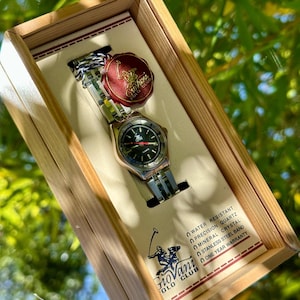 May include: A two-tone watch with a black dial and a gold-colored bezel, presented in a wooden box. The watch features a stainless steel band and a red and gold tag. The watch is water resistant and has a one-year warranty.