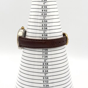 May include: A wristwatch with a brown leather band is displayed on a white and black sizing cone. The watch has a gold-colored bezel and a silver-colored face. The cone is marked with inch measurements from 2 3/4" to 10 3/4".