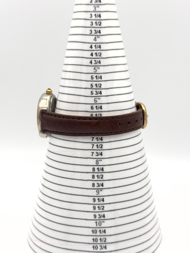 May include: A wristwatch with a brown leather band is displayed on a white and black sizing cone. The watch has a gold-colored bezel and a silver-colored face. The cone is marked with inch measurements from 2 3/4" to 10 3/4".