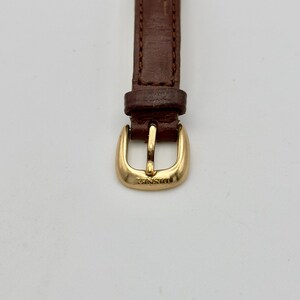 May include: Close-up of a watch strap with a brown leather band and a gold-colored buckle. The buckle is rectangular with rounded edges and the word "Disney" is engraved on it.