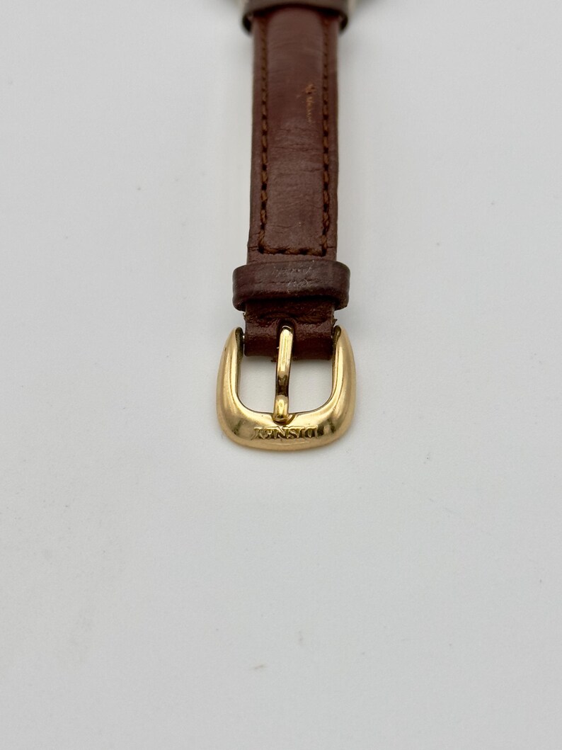 May include: Close-up of a watch strap with a brown leather band and a gold-colored buckle. The buckle is rectangular with rounded edges and the word "Disney" is engraved on it.