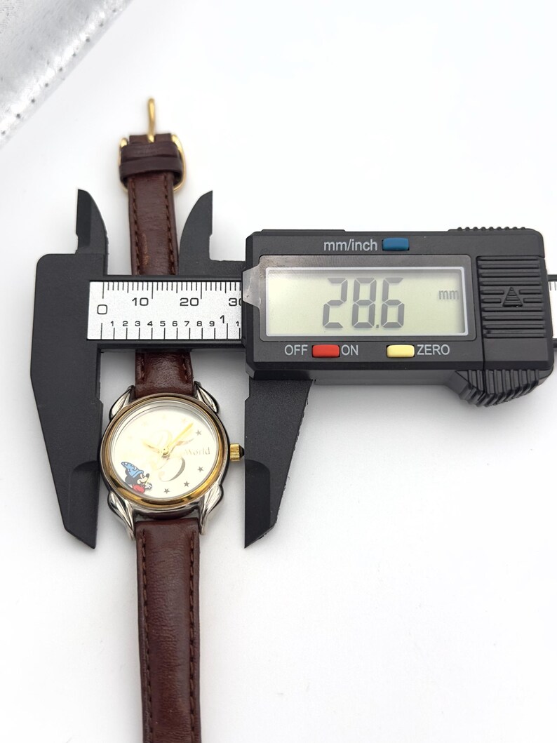 May include: A vintage wristwatch with a brown leather band and a gold-toned bezel. The watch face features a Mickey Mouse design. The digital caliper shows the watch is 28.6 mm wide. The watch is being measured against a white background.