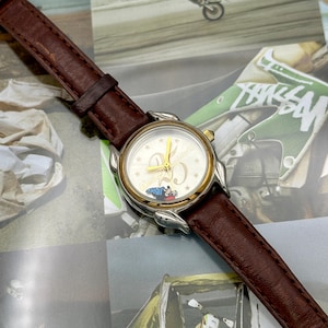 May include: A wristwatch with a brown leather band and a gold and silver-toned case. The watch face is white with gold hands and the text "Disney World 25". A small image of Mickey Mouse is visible on the watch face.