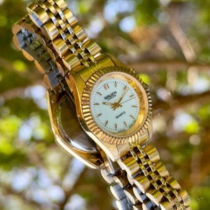 Pode incluir: Um relógio de quartzo Gruen Everlite dourado com mostrador branco e ponteiros dourados. O relógio tem uma moldura dourada e uma pulseira de elos dourados. O relógio é exibido em uma superfície reflexiva.