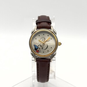 May include: A silver and gold-toned wristwatch with a brown leather band. The watch face features Mickey Mouse in a wizard hat, the number 25, and the words "Walt Disney World". The watch is displayed on a clear stand.