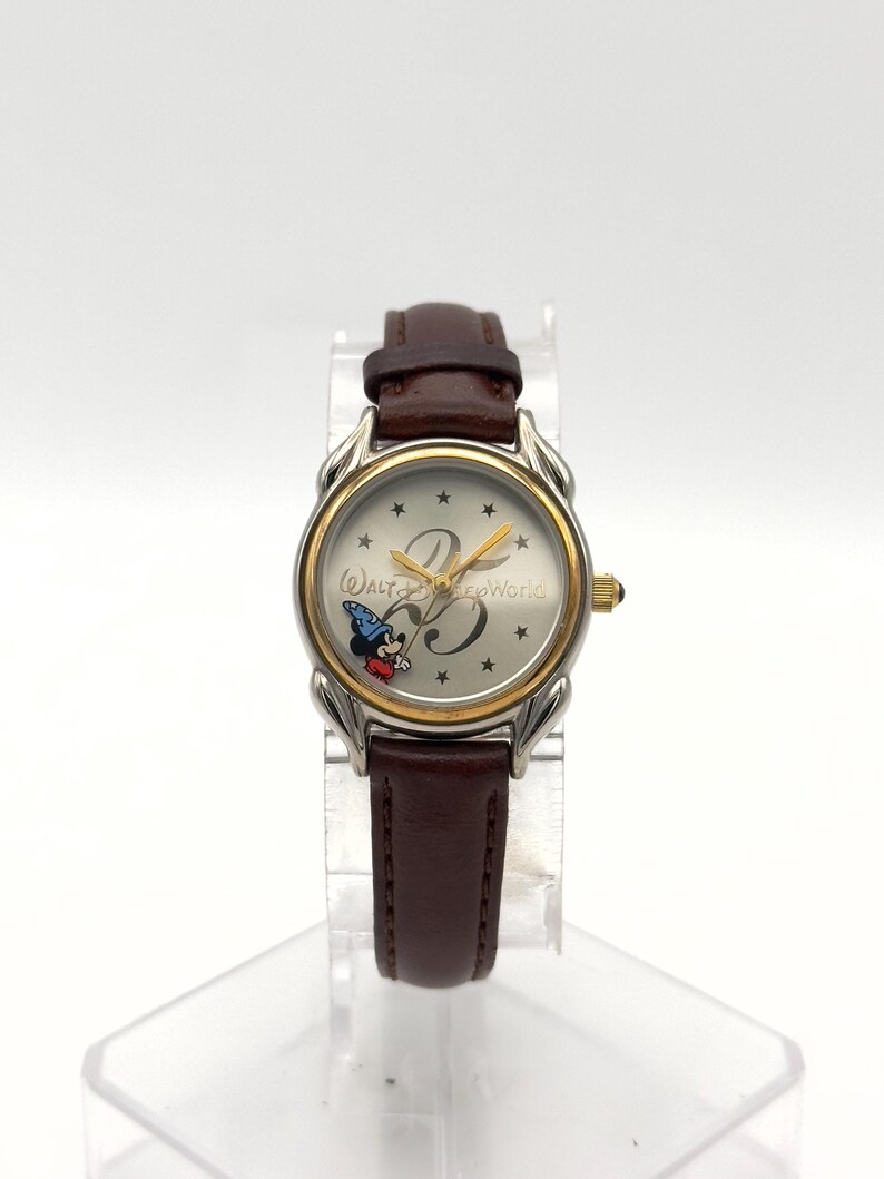 May include: A silver and gold-toned wristwatch with a brown leather band. The watch face features Mickey Mouse in a wizard hat, the number 25, and the words "Walt Disney World". The watch is displayed on a clear stand.