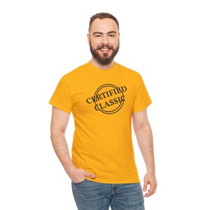 May include: Mustard yellow t-shirt with the words "CERTIFIED CLASSIC" printed in a black circular design. The t-shirt is short-sleeved and made of a soft material. The person is wearing blue jeans.