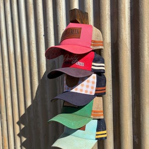 May include: A wooden hat rack displaying six baseball caps in various colours, including pink, red, green, and blue. Each cap features a mesh back and a curved brim. The caps are attached to a wooden post, mounted on a corrugated metal wall.