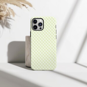 Puede incluir: Funda de teléfono verde claro con un patrón de cuadros. La funda tiene un recorte negro para la cámara y botones laterales negros. La funda está sobre una superficie blanca.