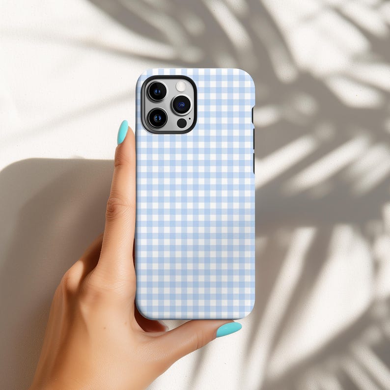 May include: A light blue and white gingham pattern phone case. The case has a square camera cutout and a black border. The phone case is held by a hand with blue nail polish.