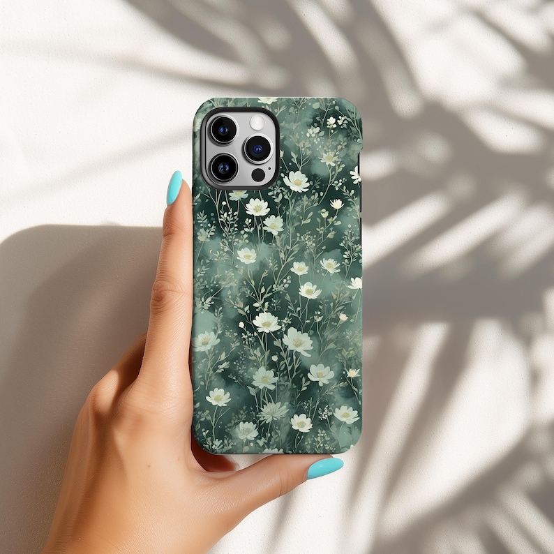 Op de afbeelding: Een smartphonehoesje met een bloemenpatroon. De hoes heeft een donkergroene achtergrond met witte bloemen en groen blad. De telefoonhoes wordt vastgehouden door een persoon met blauwe nagellak.