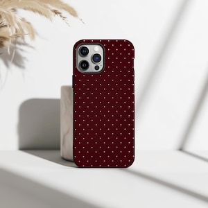 May include: A burgundy phone case with a repeating pattern of small white dots. The case has a black border and a camera cutout. The phone case is standing upright on a white surface.