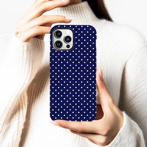 Puede incluir: Una funda de teléfono azul marino con un patrón de lunares blancos. La funda se sostiene frente a un suéter blanco. La funda del teléfono tiene un recorte para la cámara y un borde negro.