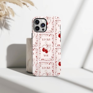 May include: A white phone case with a repeating pattern of red playing card designs. Each card features a pair of cherries, a bow, and the words "LUCKY YOU". The case has a glossy finish and a camera cutout.