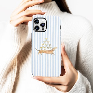 May include: A phone case with a light blue and white striped background. The case features a watercolor illustration of a dachshund dog carrying a pyramid of champagne glasses. The case is held by a person.