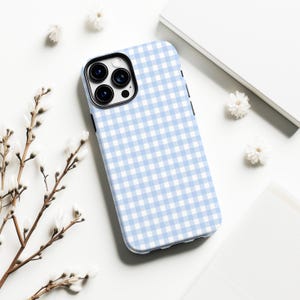 May include: A light blue and white gingham pattern phone case. The case is on a white surface with small white flowers and a sprig of a plant. The phone case has a black camera lens area.