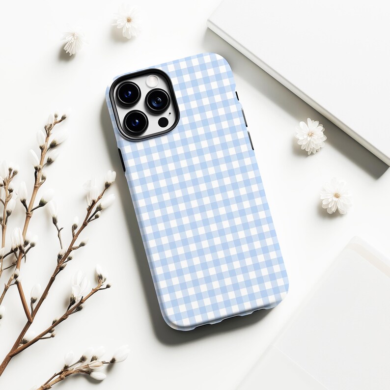 May include: A light blue and white gingham pattern phone case. The case is on a white surface with small white flowers and a sprig of a plant. The phone case has a black camera lens area.