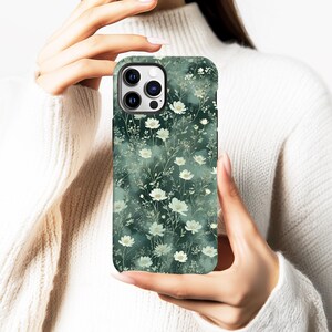 Op de afbeelding: Een smartphonehoesje met een bloemenpatroon. De hoes heeft een donkergroene achtergrond met een patroon van witte bloemen en groene bladeren. De hoes wordt in iemands handen gehouden.