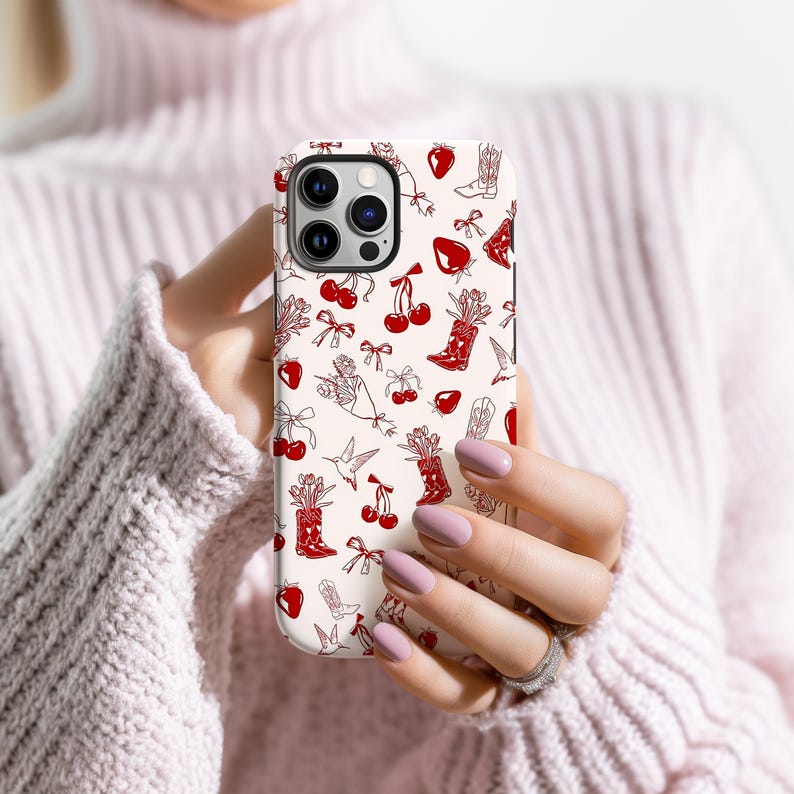 Pu&ograve; includere: Una custodia per telefono bianca con un motivo ripetuto di ciliegie rosse, stivali da cowboy, fragole, fiocchi e bouquet floreali. La custodia &egrave; tenuta tra le mani di una persona.