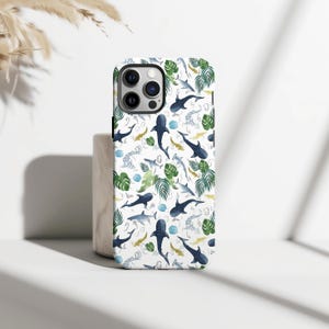 May include: A white phone case with a repeating pattern of watercolour sharks, tropical leaves, and gold fish. The case has a glossy finish and a camera cutout. The design includes blue, green, and gold accents.