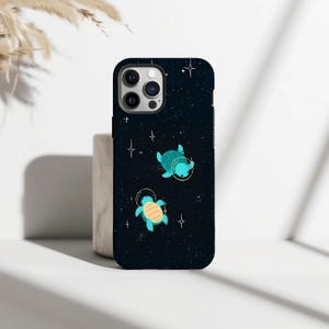 May include: A black phone case with a space-themed design. It features two cartoon turtles in astronaut helmets, rendered in teal and yellow, against a starry night sky. White stars and dots are scattered across the case, creating a cosmic effect.