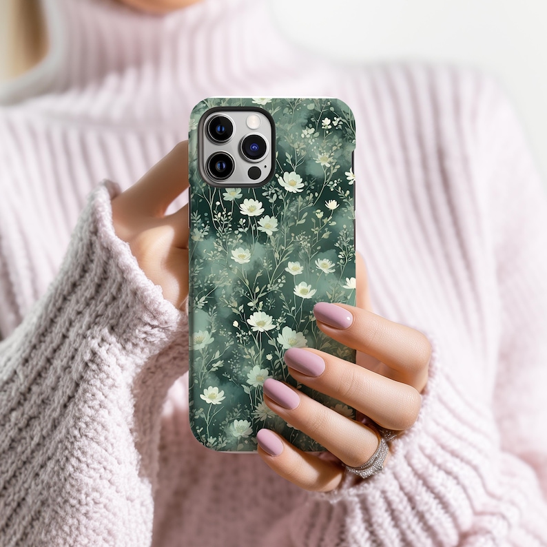 Op de afbeelding: Een telefoonhoesje met een bloemenpatroon. De hoes heeft een donkergroene achtergrond met witte bloemen en groene bladeren. De hoes heeft een glanzende afwerking en is ontworpen voor een smartphone.
