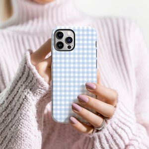May include: A smartphone case with a light blue and white gingham pattern. The case has a black camera module and a smooth, glossy finish. The case is held in front of a pink sweater.