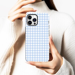 May include: A light blue and white gingham pattern phone case. The case has a black camera cutout and is held by a person wearing a white sweater. The case is designed for a smartphone.