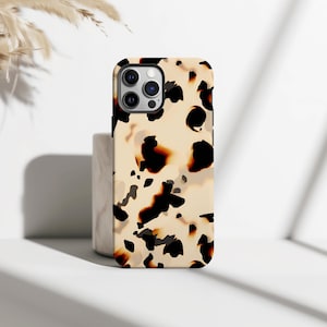 Può includere: Una custodia per telefono con motivo tartarugato. La custodia ha uno sfondo color crema con macchie irregolari marrone scuro e nere. La custodia del telefono è rettangolare con angoli arrotondati e un ritaglio per la fotocamera.