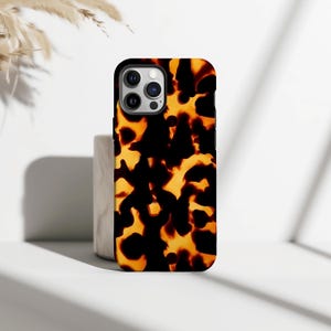 Puede incluir: Una funda de teléfono inteligente con un patrón de carey en tonos negros y ámbar. La funda tiene un acabado brillante y un recorte para la cámara. La funda del teléfono se muestra sobre una superficie blanca.