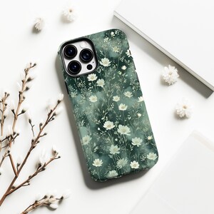 Op de afbeelding: Een smartphonehoesje met een bloemenpatroon. De hoes heeft een donkergroene achtergrond versierd met witte bloemen en slanke groene stelen. Het ligt op een wit oppervlak, aangevuld met extra bloemelementen.