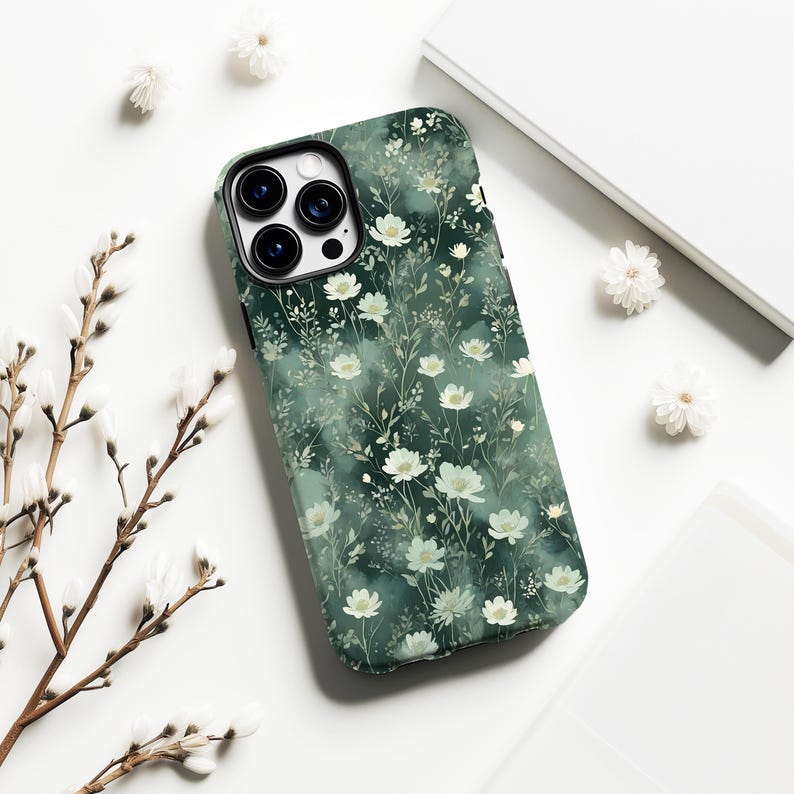 Op de afbeelding: Een smartphonehoesje met een bloemenpatroon. De hoes heeft een donkergroene achtergrond versierd met witte bloemen en slanke groene stelen. Het ligt op een wit oppervlak, aangevuld met extra bloemelementen.