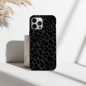 Puede incluir: Una funda de teléfono negra con un diseño de estampado de leopardo gris oscuro. La funda está hecha de un material duradero y tiene un aspecto elegante y moderno. La funda está diseñada para proteger el teléfono de arañazos y daños.